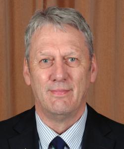 A middle-aged man with short gray hair, wearing a dark suit, blue and white striped shirt, and a dark tie, poses in front of a plain brown curtain background.