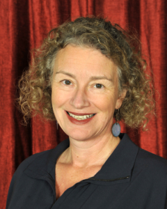 A woman with curly hair, wearing a dark top and blue earrings, smiles in front of a red curtain backdrop.