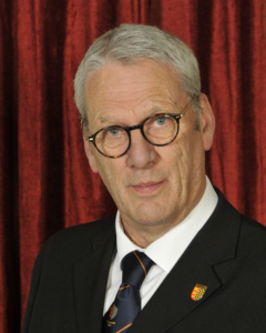 An older man with gray hair and glasses wears a black suit and tie, posing in front of a red curtain background.