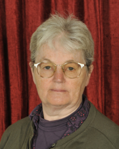 An older woman with short gray hair, glasses, and a neutral expression stands in front of a red curtain, wearing a green sweater and a purple shirt.