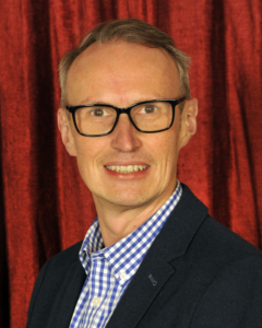 A man with short blond hair and glasses wearing a blue blazer and checkered shirt stands in front of a red curtain.