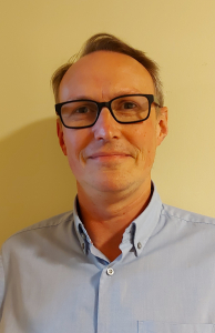 A middle-aged man with short light brown hair and glasses, wearing a light blue button-up shirt, stands in front of a plain beige wall, looking at the camera with a neutral expression.
