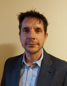 A man with short brown hair and stubble, wearing a blue shirt and dark blazer, stands against a plain beige background, looking directly at the camera with a neutral expression.