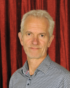 An older man with short gray hair and a patterned shirt stands in front of a red curtain, looking at the camera with a neutral expression.