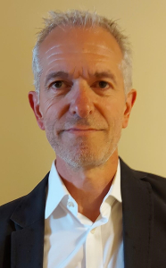 A middle-aged man with short gray hair and stubble, wearing a white dress shirt and a dark blazer, stands against a plain beige background, looking directly at the camera with a neutral expression.