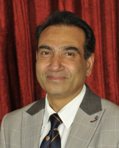 A man in a light-colored suit, white shirt, and patterned tie poses in front of a red curtain background.