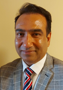 A man with dark hair, wearing a gray plaid suit jacket, white shirt, and a red, white, and blue striped tie, smiles slightly while standing against a plain beige background.