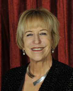 Smiling older woman with short blonde hair, wearing earrings and a silver necklace, poses in front of a red curtain background.