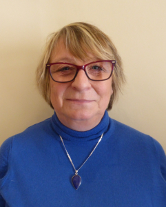 A woman with short, light brown hair wears red glasses, a blue turtleneck sweater, and a silver necklace with a blue pendant, standing in front of a plain beige wall.