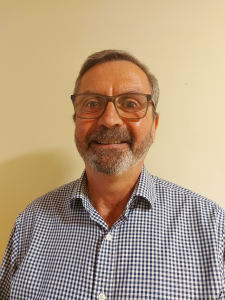 A smiling older man with glasses, gray hair, and a beard, wearing a blue and white checkered shirt, stands in front of a plain light-colored wall.