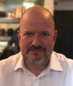 A middle-aged man with a shaved head and a short gray beard wearing a white collared shirt, sitting indoors with a blurred background of shelves and another person.