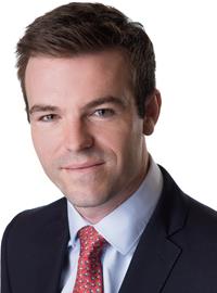 A man with short brown hair wearing a dark suit, light blue shirt, and red patterned tie, smiling slightly, posed against a white background.