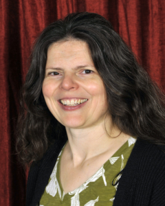 A woman with long, wavy brown hair smiles at the camera, wearing a green patterned blouse and black cardigan, in front of a red curtain background.