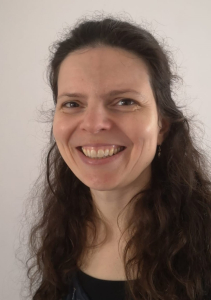 A woman with long, wavy brown hair and light skin smiles at the camera against a plain light background. She is wearing a dark top and small dangling earrings.