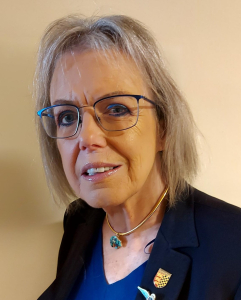 An older woman with gray hair and glasses is wearing a dark blazer, a blue top, and a gold necklace with a turquoise pendant. She has two pins on her blazer and is standing in front of a neutral beige background.