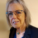 An older woman with gray hair and glasses is wearing a dark blazer, a blue top, and a gold necklace with a turquoise pendant. She has two pins on her blazer and is standing in front of a neutral beige background.