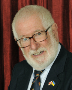 Elderly man with white hair and beard, wearing glasses, a suit, and a tie, standing in front of a red curtain. He has pins of the British and Ukrainian flags on his lapel.