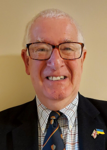 An older man with short white hair and glasses smiles at the camera. He is wearing a dark suit, a patterned tie, and a lapel pin featuring the UK and Ukrainian flags.