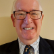 An older man with short white hair and glasses smiles at the camera. He is wearing a dark suit, a patterned tie, and a lapel pin featuring the UK and Ukrainian flags.