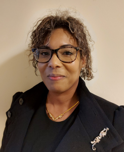 A woman with curly brown hair, wearing glasses, a black coat with a silver leopard brooch, and a gold necklace, stands against a plain light-colored wall, smiling slightly at the camera.