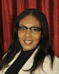 A woman with long dark hair, glasses, and a light-colored blazer smiles in front of a red curtain background.