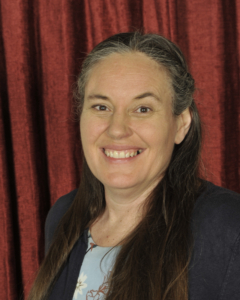 A woman with long brown hair and a blue blazer smiles in front of a red curtain backdrop.