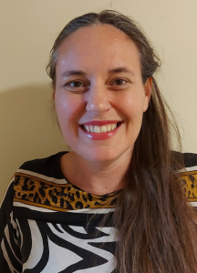 A woman with long brown hair, smiling at the camera, wearing a patterned top with black, white, and yellow designs, standing against a plain light-colored background.