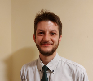 A man with short brown hair and a beard, wearing a white collared shirt and a green tie, smiles at the camera against a plain beige wall.