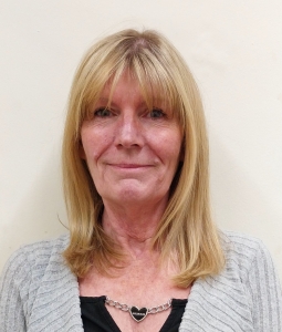 A woman with straight, shoulder-length blonde hair wearing a gray sweater, black top, and a heart-shaped necklace stands against a plain light-colored wall.