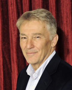 A middle-aged man with short gray hair wearing a dark jacket and light shirt, posing in front of a red velvet curtain.