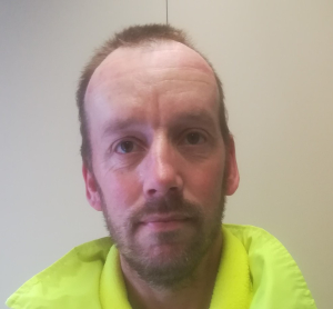 A man with light skin, short brown hair, and a beard looks at the camera. He is wearing a neon yellow jacket and standing in front of a plain, light-colored wall.