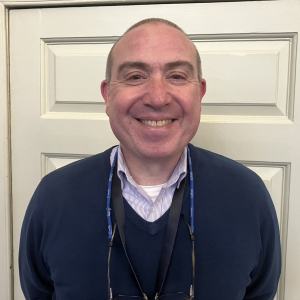 A man with short hair smiles at the camera. He is wearing a navy sweater over a collared shirt, with glasses hanging from a lanyard around his neck, standing in front of a cream-colored paneled door.