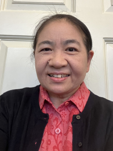 A woman with dark hair pulled back smiles at the camera. She is wearing a red patterned shirt under a black cardigan, standing in front of a light-colored paneled door.