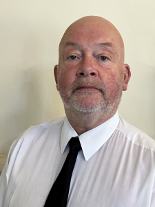 A middle-aged, bald man with a trimmed gray beard wearing a white shirt and black tie, sitting against a plain, light-colored background.