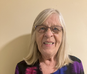 An older woman with long gray hair and glasses smiles at the camera while standing against a beige wall. She is wearing a black top with purple and blue patterns.