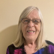 An older woman with long gray hair and glasses smiles at the camera while standing against a beige wall. She is wearing a black top with purple and blue patterns.