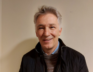 A smiling older man with gray hair wearing a black quilted jacket and a brown patterned sweater stands against a plain light-colored wall.