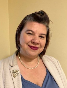 A woman with short, dark hair styled to the side is smiling. She wears a light blazer, a blue top, a pearl necklace, and a brooch, standing against a beige wall.