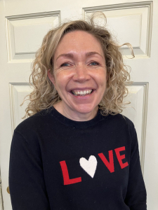 A woman with curly blonde hair smiles at the camera, wearing a black sweater with the word "LOVE" in red and white letters, standing in front of a white door.