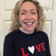 A woman with curly blonde hair smiles at the camera, wearing a black sweater with the word "LOVE" in red and white letters, standing in front of a white door.