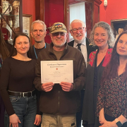 A group of seven adults pose indoors, smiling. The man in the center wears a cap and glasses and holds a Certificate of Appreciation. Shelves and framed items decorate the warm, red-toned room behind them.