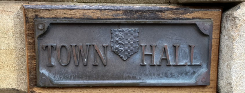 A metal sign mounted on a stone wall reads "TOWN HALL" with a decorative crest or shield between the two words. The plaque has a weathered appearance.