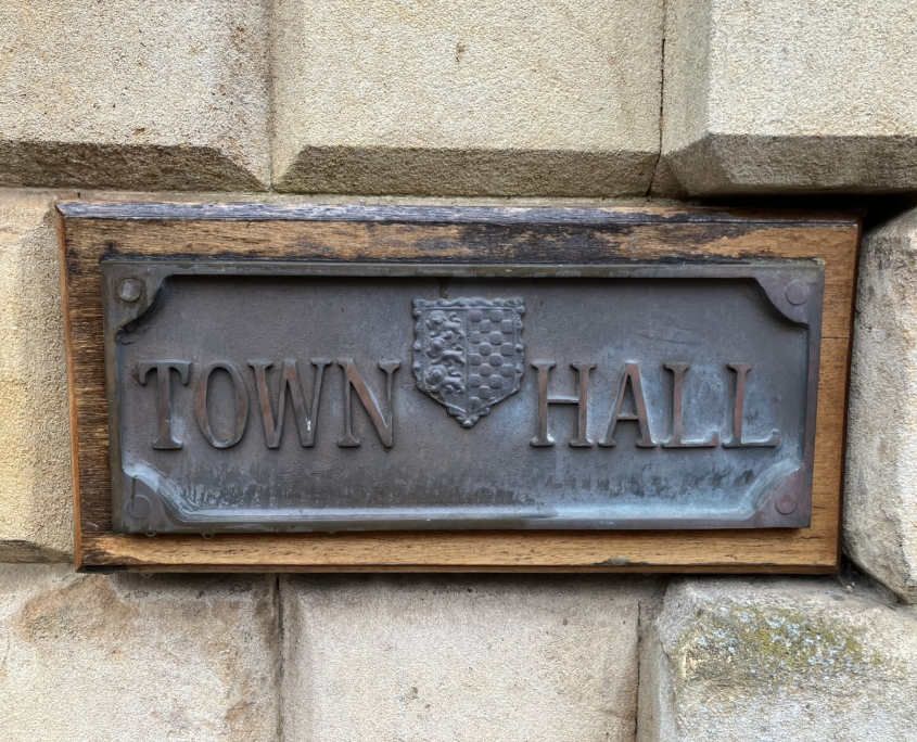 A metal sign mounted on a stone wall reads "TOWN HALL" with a decorative crest or shield between the two words. The plaque has a weathered appearance.