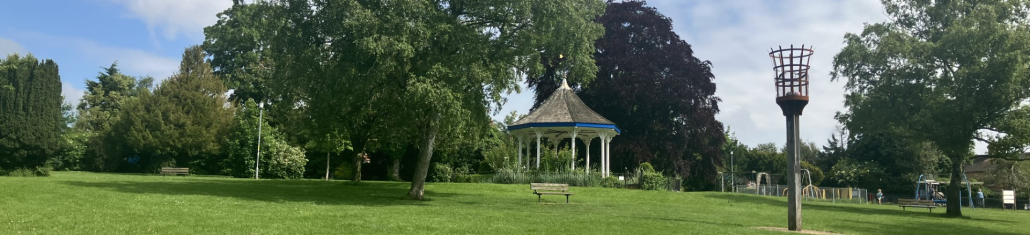 A green park with scattered trees, benches, a white gazebo with a blue roof, a metal torch-like structure, and a playground in the background under a partly cloudy sky.
