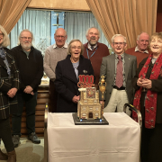 A group of nine smiling older adults stand behind a decorated cake celebrating 1000 organ recitals, with curtains and an organ visible in the background.