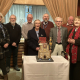 A group of nine smiling older adults stand behind a decorated cake celebrating 1000 organ recitals, with curtains and an organ visible in the background.
