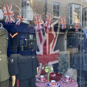 AGE UK shop window display with military uniforms on mannequins, Union Jack flags, bunting, and a table set with teacups and flowers.