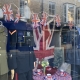 AGE UK shop window display with military uniforms on mannequins, Union Jack flags, bunting, and a table set with teacups and flowers.