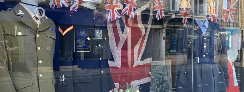 AGE UK shop window display with military uniforms on mannequins, Union Jack flags, bunting, and a table set with teacups and flowers.