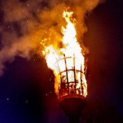 A large beacon fire burns atop a tall metal basket at night, with a crowd of people and trees silhouetted in the background.
